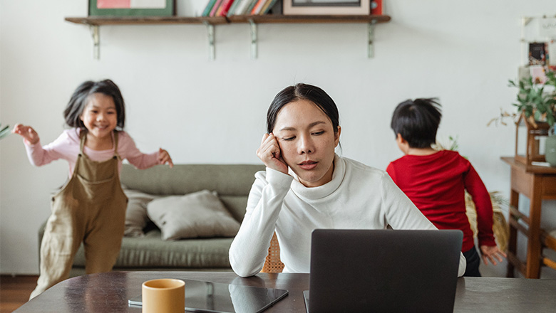 Exhausted woman working from home in front of her laptop with her two children in the background running to show how remote work establishes equality at the workplace